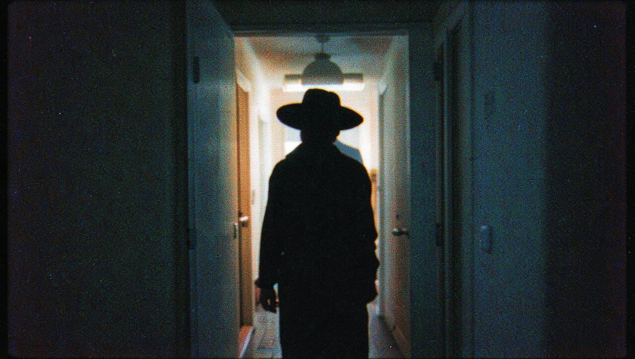 A silhouette in a wide-brimmed hat, standing in a bedroom doorway, backlit by a dim hallway. The most reported composition in the file.