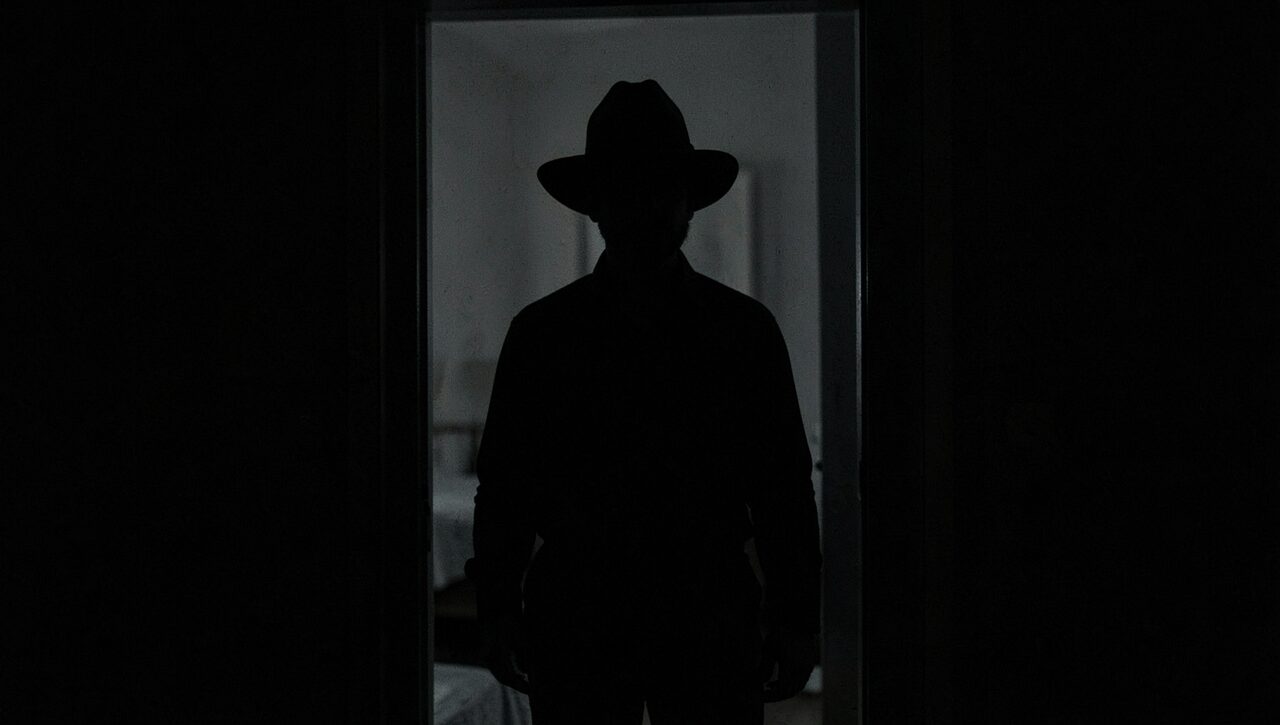 A figure in a wide-brimmed hat, centered in a doorway, slightly closer than is comfortable. Both rooms dark. He is simply there.