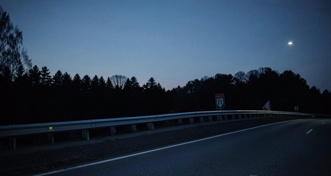 A metal guardrail along a dark road, tree line silhouetted beyond. One witness said the thing stepped over it.