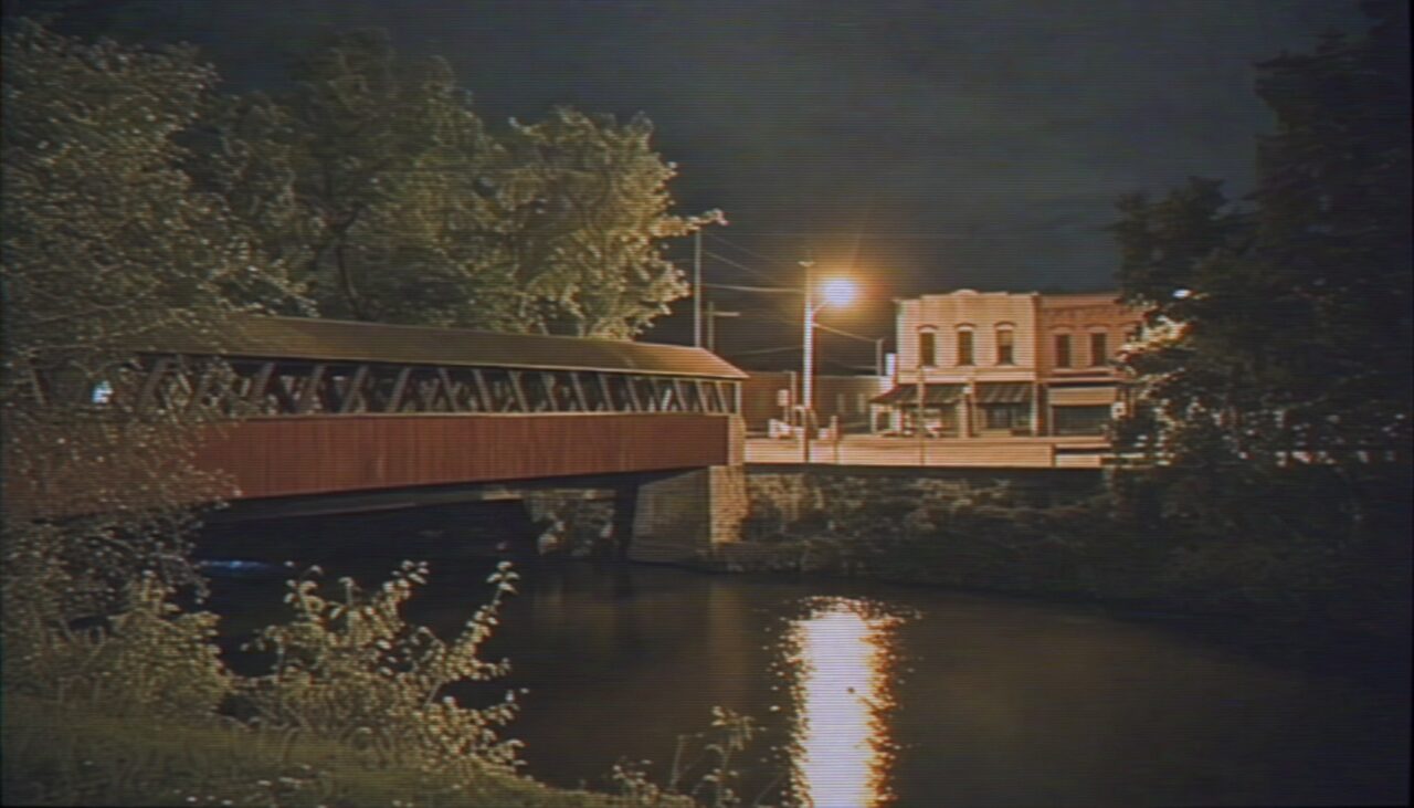 Loveland at night, seen from across the river — covered bridge, closed storefronts, one streetlight doing the work of several.