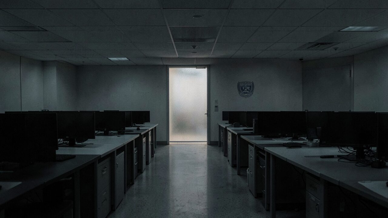 A secure Pentagon office corridor late at night. Empty desks, dark monitors, one frosted door still lit at the far end.